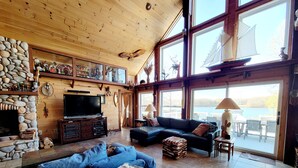 Un interior de cabaña acogedor, con chimenea de piedra, techo de madera, ventanas grandes y un televisor.