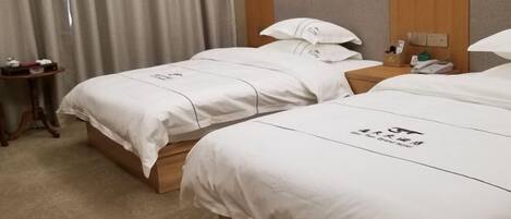 A hotel room with two beds, each with white bedding and a logo on the headboard. There is a wooden nightstand with a phone and a small table with a lamp and a bottle.