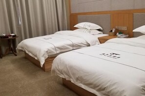Ein Hotelzimmer mit zwei Betten, jeweils mit weißer Bettwäsche und einem Logo an der Kopfteil. Es gibt einen hölzernen Nachttisch mit einem Telefon und einen kleinen Tisch mit einer Lampe und einer Flasche.