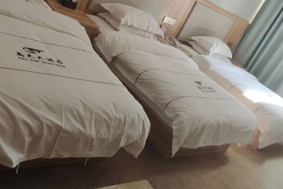 Une chambre d’hôtel avec trois lits, chacun recouvert de draps blancs. Les lits sont disposés en rangée et dotés de têtes de lit en bois.