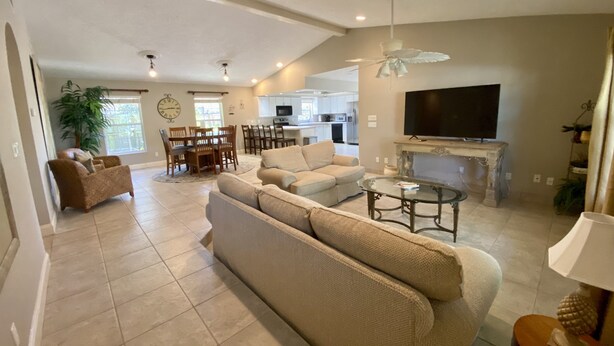 A spacious living room with a large sofa, a dining area with wooden chairs, a kitchen in the background, and a TV mounted above a fireplace. - Vanderbilt Beach Retreat- 4 Bed 1-level, Spacious, & Pet-Friendly - 3 King Beds (Naples)
