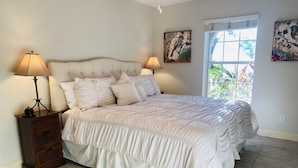 A bedroom with a large bed, two bedside lamps, a nightstand, and a window with a view of greenery. - Vanderbilt Beach Retreat- 4 Bed 1-level, Spacious, & Pet-Friendly - 3 King Beds (Naples)