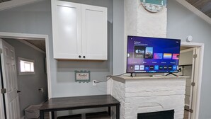 Una sala de estar con chimenea de ladrillo blanco, un televisor de pantalla plana montado arriba y una pequeña mesa con dos sillas.