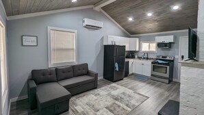Cocina compacta con horno empotrado, microondas y refrigerador. Un sofá gris y una mesita pequeña se encuentran en la zona de estar.