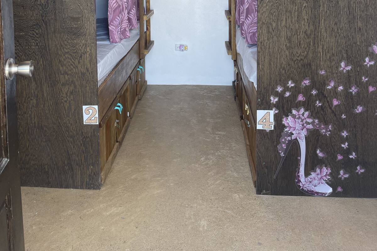 Um corredor estreito com beliches, portas numeradas e um painel floral parcial na parede.