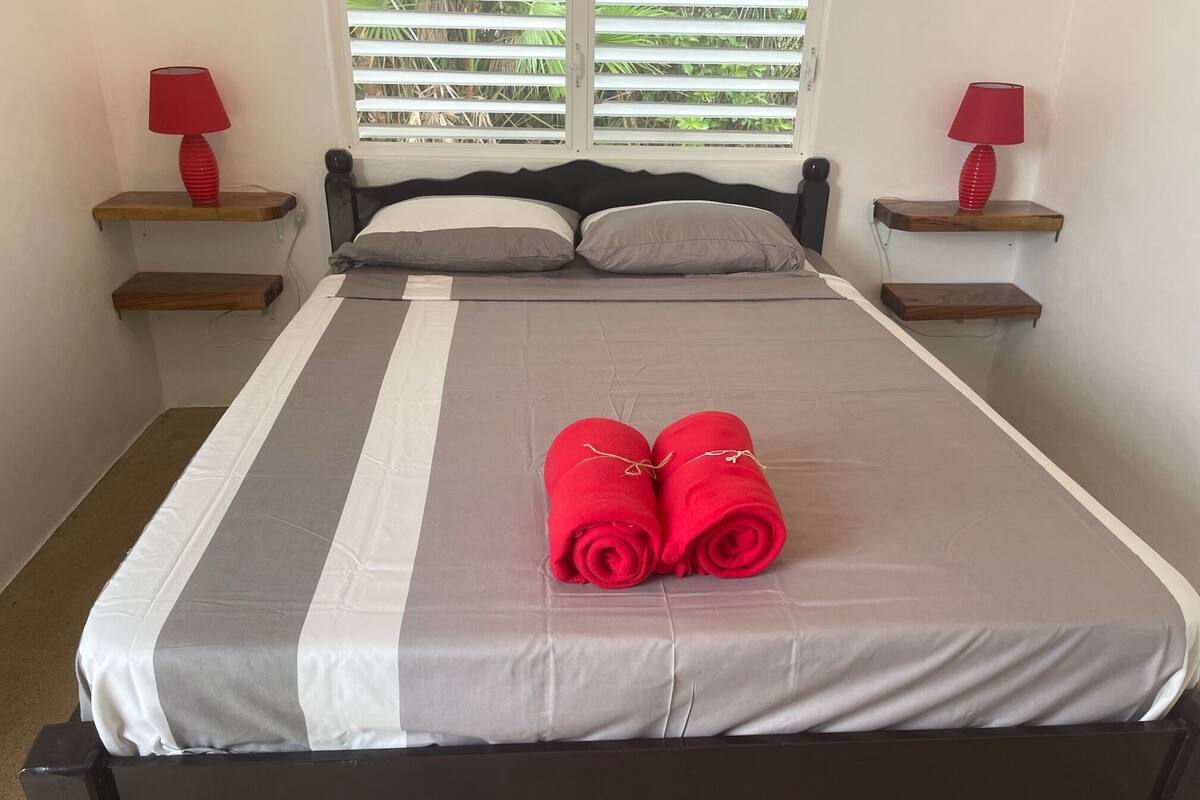 Cama bem arrumada com colcha listrada em cinza e branco, dois travesseiros e duas toalhas vermelhas sobre a cama. Há duas mesas de cabeceira com luminárias vermelhas e uma janela com persianas horizontais.