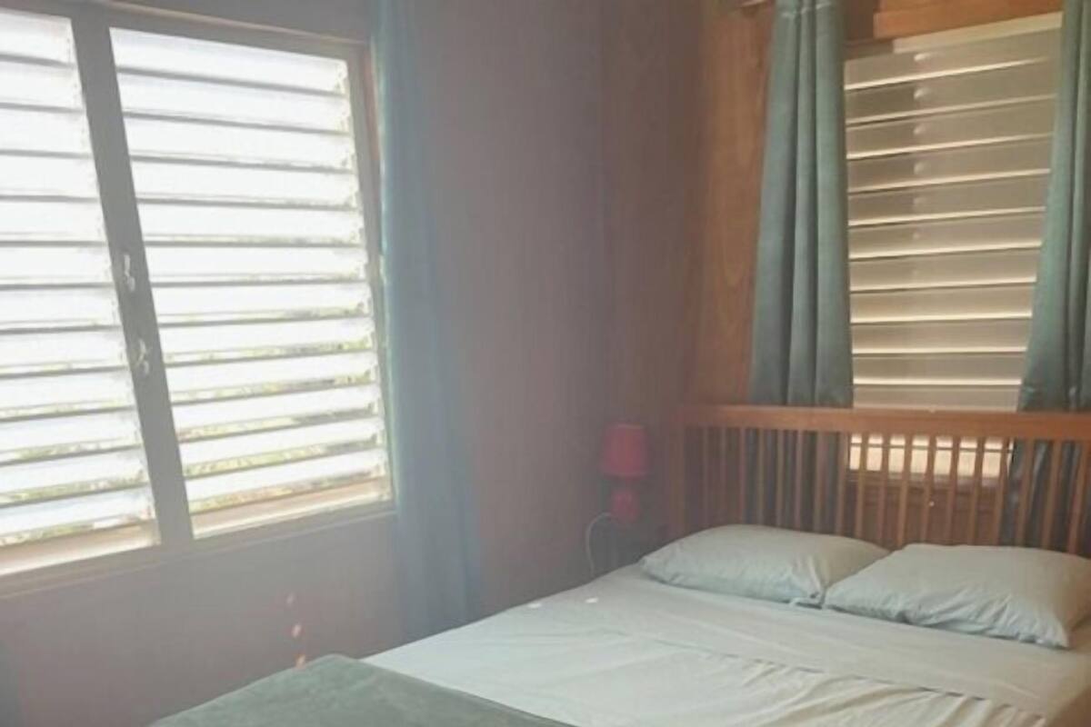 Uma cama de solteiro com estrutura de madeira e colcha verde num quarto com piso de madeira e janela com persianas.
