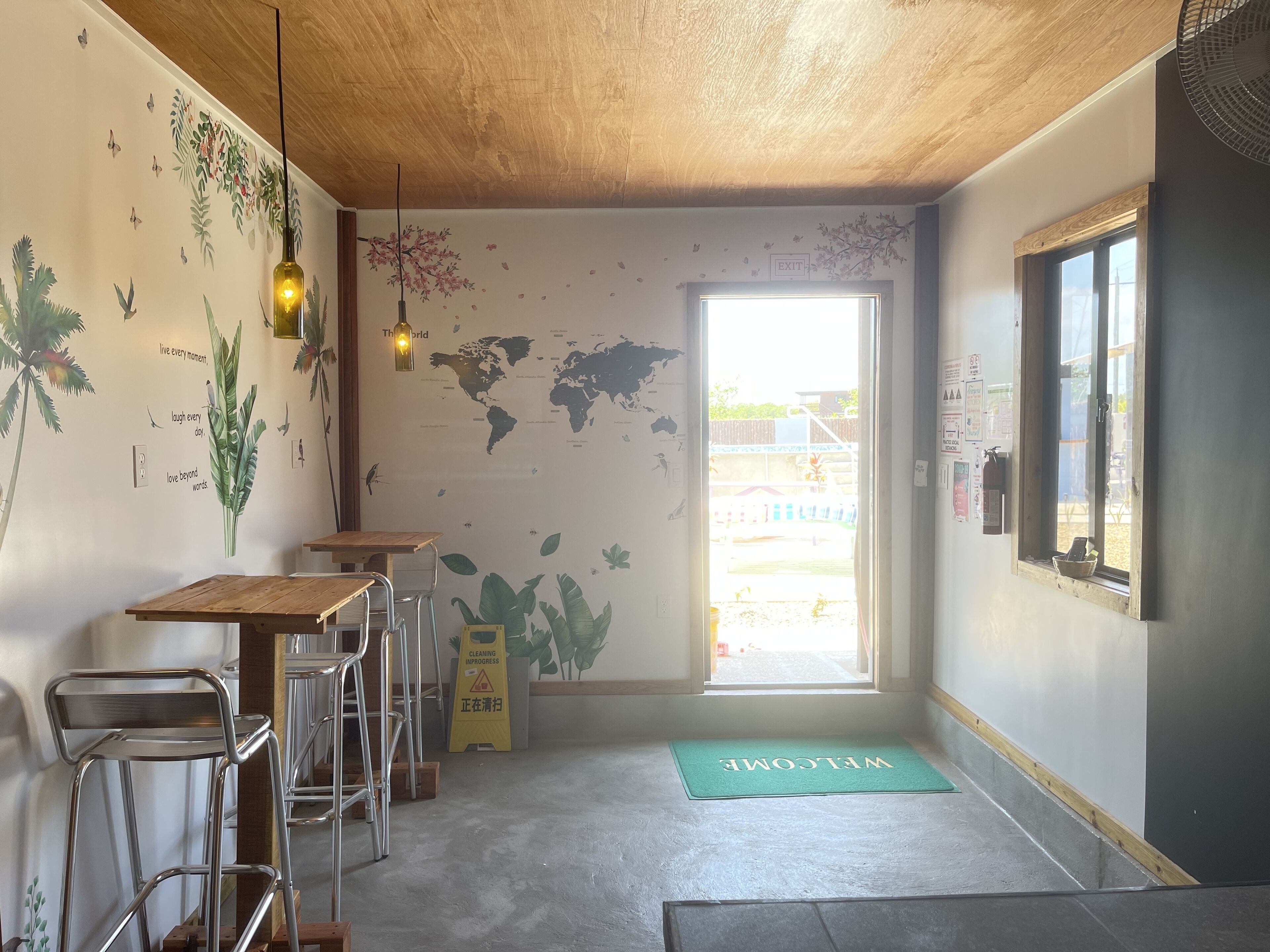A cafe interior with a wooden ceiling, world map wall art, a 'WELCOME' mat, and a 'CLEANING INPROGRESS' sign.