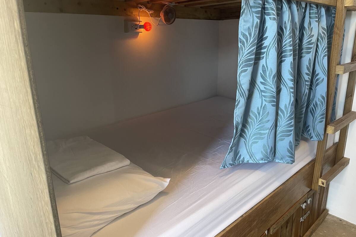 Uma cama beliche com um colchão simples, cabeceira de madeira e uma pequena mesa de cabeceira.