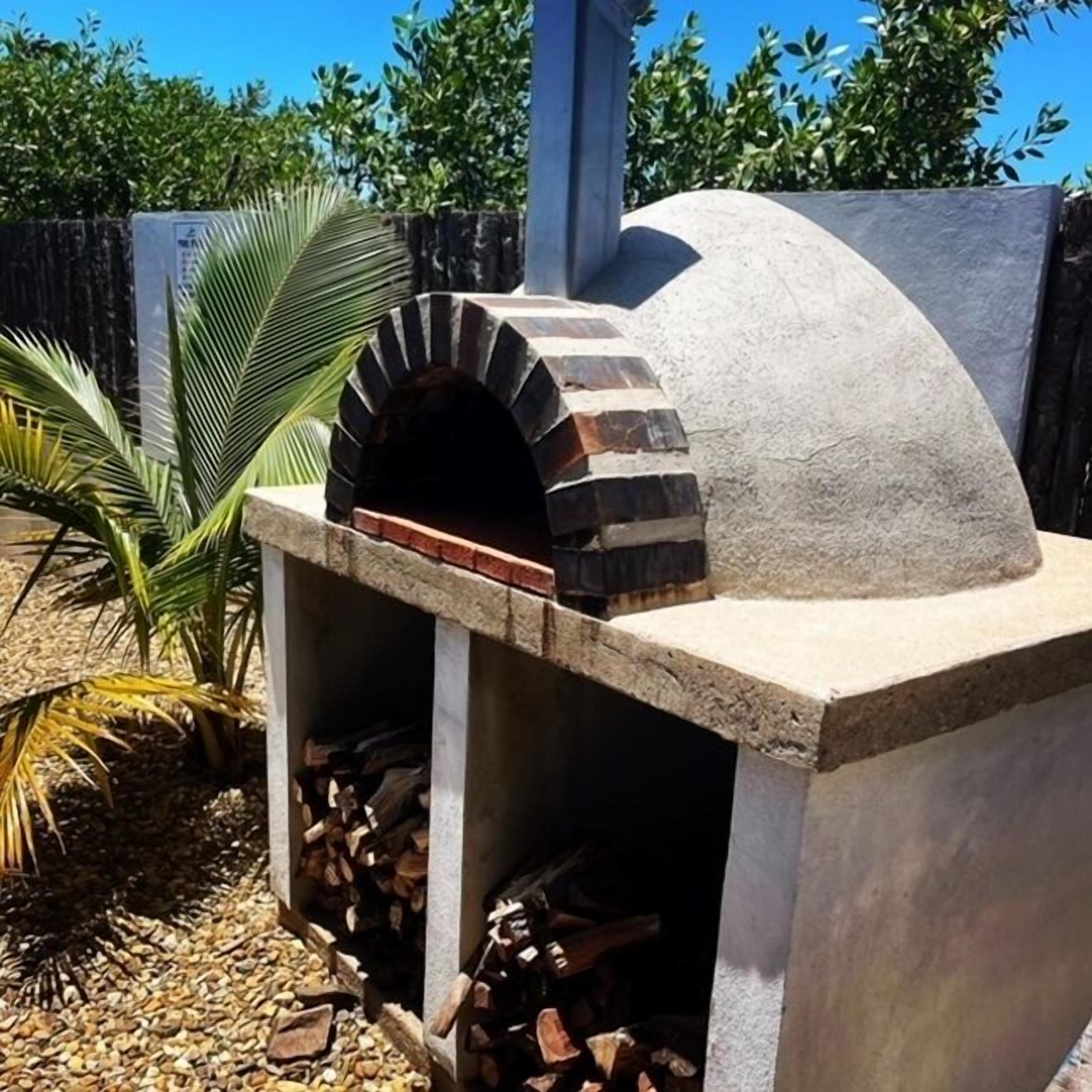 Un horno de pizza de piedra con chimenea de ladrillo, rodeado por una base de concreto y un pequeño jardín con plantas de palma.
