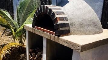 Un horno de pizza de piedra con chimenea de ladrillo, rodeado por una base de concreto y un pequeño jardín con plantas de palma.