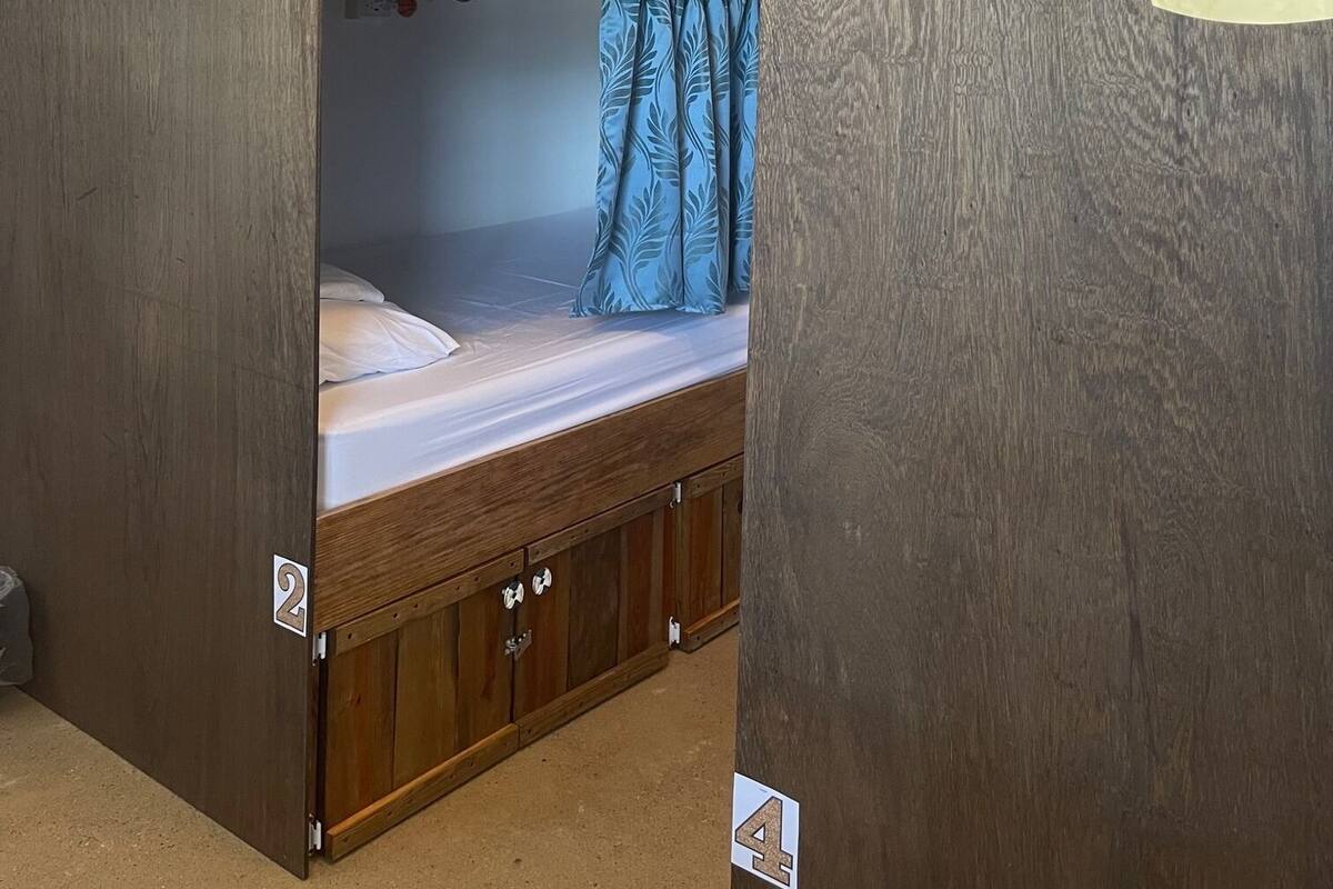 Quarto com beliche, ventilador de teto, colchões de madeira e parede decorada com motivos de coroa e peixes.