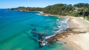 A coastal beach with clear turquoise waters, rocky shoreline, and a forested hillside in the background.