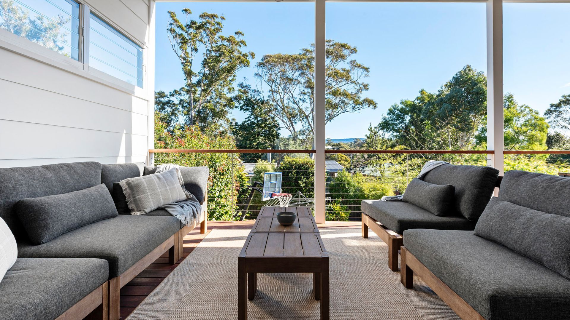 Un espace extérieur moderne, aménagé avec deux canapés gris et une table basse en bois, offrant une vue sur un jardin luxuriant et des arbres.
