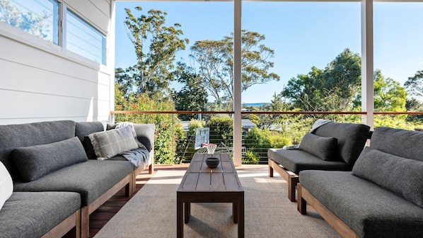 A modern outdoor seating area with two grey sofas and a wooden coffee table, overlooking a lush garden and trees.