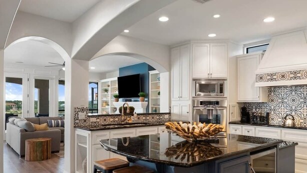 A modern kitchen with white cabinetry, a central island with stools, a flat-screen TV, and a patterned backsplash.
