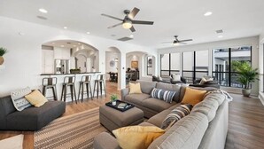 A spacious living area with a sofa, ottoman, and a coffee table. There are ceiling fans, a kitchen in the background, and large windows.