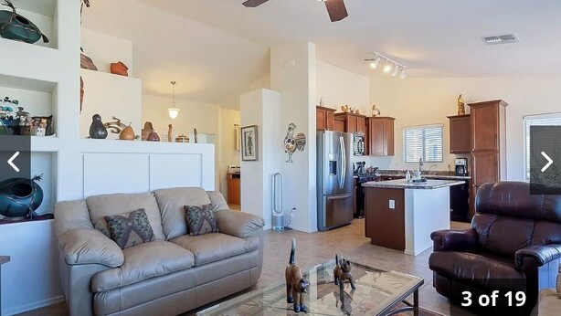 A spacious living area with a sofa, a glass coffee table, and a leather armchair. A kitchen with a refrigerator and cabinets is visible in the background.