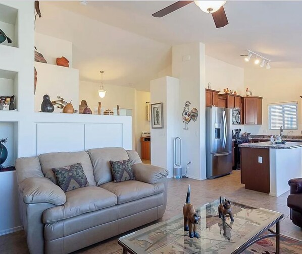 Un espacio de sala amplio con un sofá, una mesa de centro de vidrio y un sillón de cuero. Se ve una cocina con refrigerador y armarios al fondo.