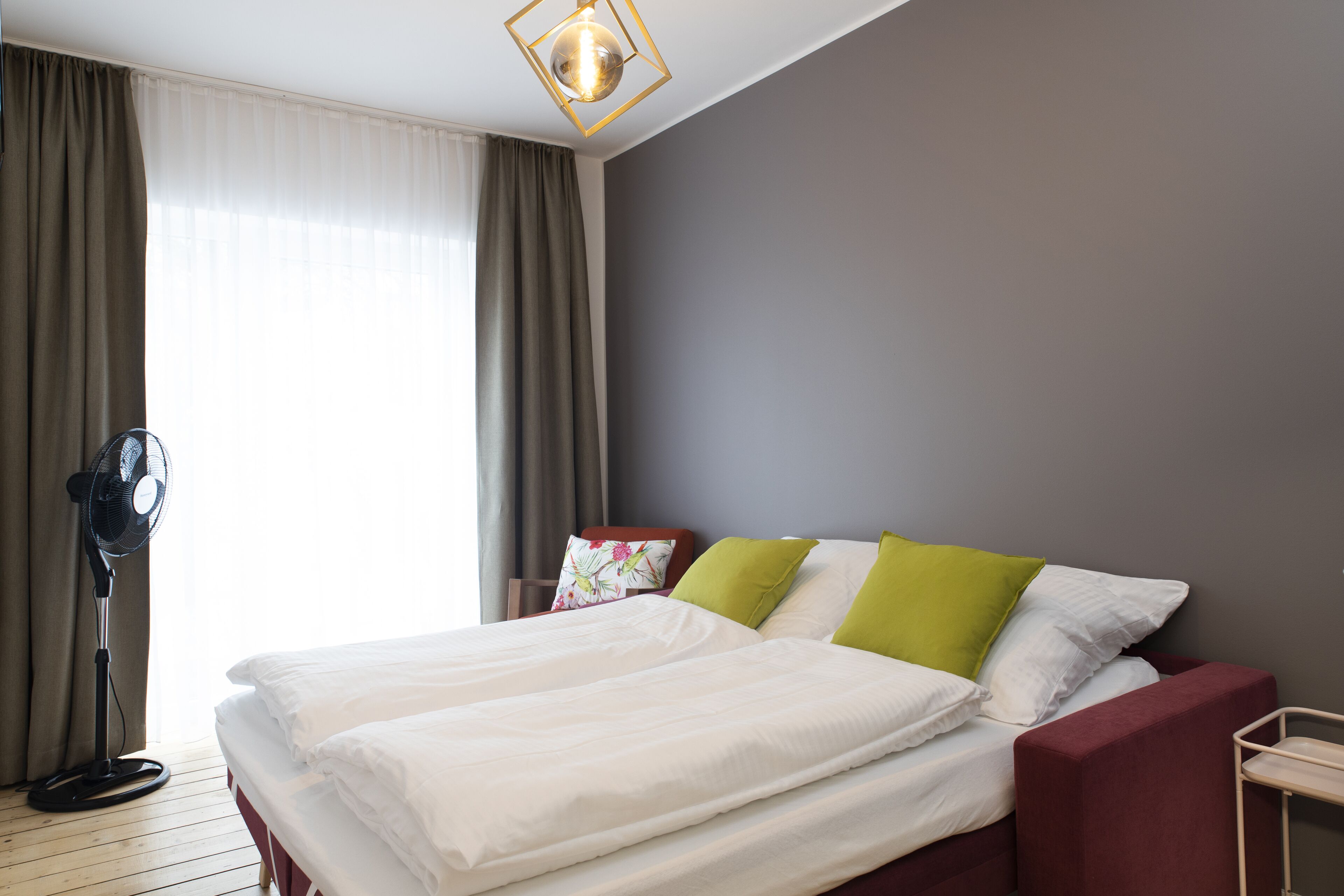 A hotel room with a bed, a standing fan, and a window with curtains.