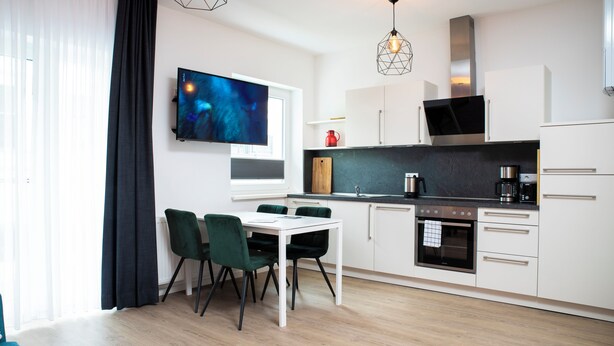 A modern kitchen with white cabinets, a dining table with green chairs, and a large flat-screen TV on the wall. - Villa am Bahnhof (Fulda)