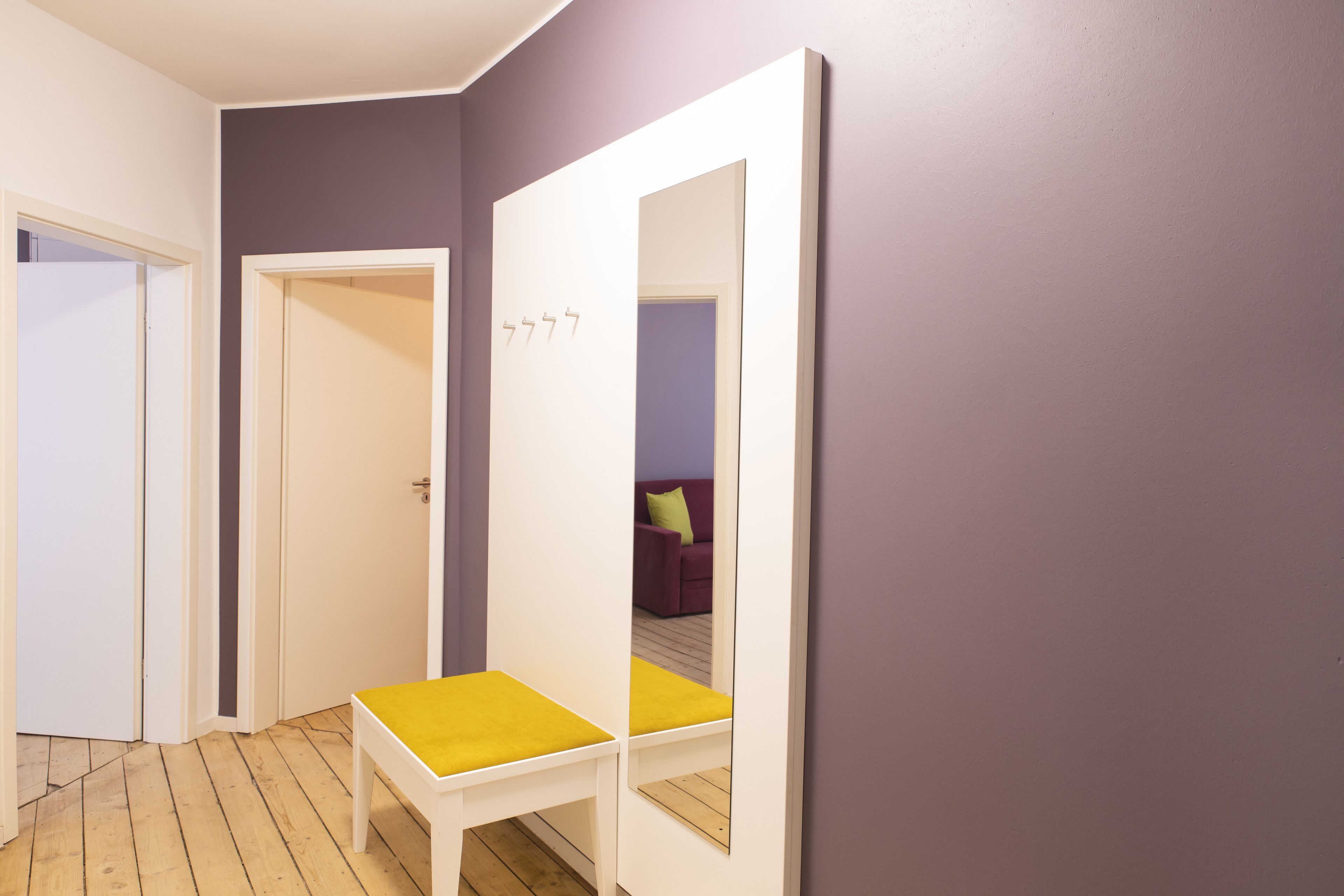 A hallway with a white bench, a door, and a large mirror reflecting a purple wall and a room with a purple sofa.