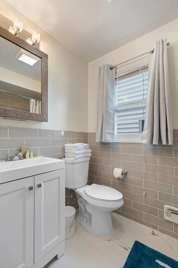 Un baño con inodoro blanco, lavamanos con espejo y ventana con cortinas.