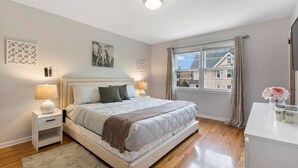 A bedroom with a bed, bedside tables, a window with curtains, and a view of buildings outside. - The Lennox Stay - Close to NYC ☆ Free Parking (Bloomfield)