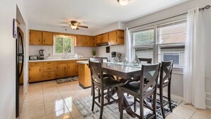 A kitchen with wooden cabinets, a dining table with chairs, a ceiling fan, and a window with curtains. - The Lennox Stay - Close to NYC ☆ Free Parking (Bloomfield)