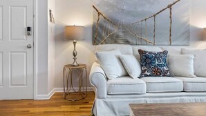 A room with a white door, a white sofa with patterned cushions, a small round table with a lamp, and a large abstract painting on the wall. - The Lennox Stay - Close to NYC ☆ Free Parking (Bloomfield)