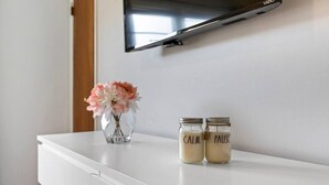 A white dresser with two jars labeled "CALM" and "PAUSE" on top, a vase with flowers, and a TV mounted on the wall. - The Lennox Stay - Close to NYC ☆ Free Parking (Bloomfield)