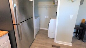 Una cocina compacta con un refrigerador de acero inoxidable, una lavadora blanca pequeña y un microondas. Hay dos macetas en la pared y una pequeña mesa con una silla.