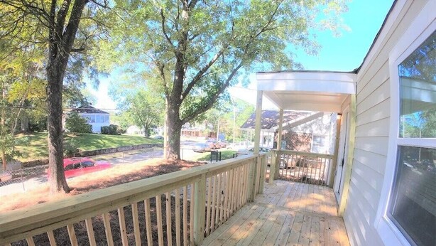 Una terraza de madera con barandilla, un porche cubierto y vista a una calle residencial con casas y árboles.