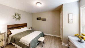 Un dormitorio con cabecera de madera, colcha verde, mesita de noche, taburete pequeño, cómoda, espejo y un adorno colgante en la pared.