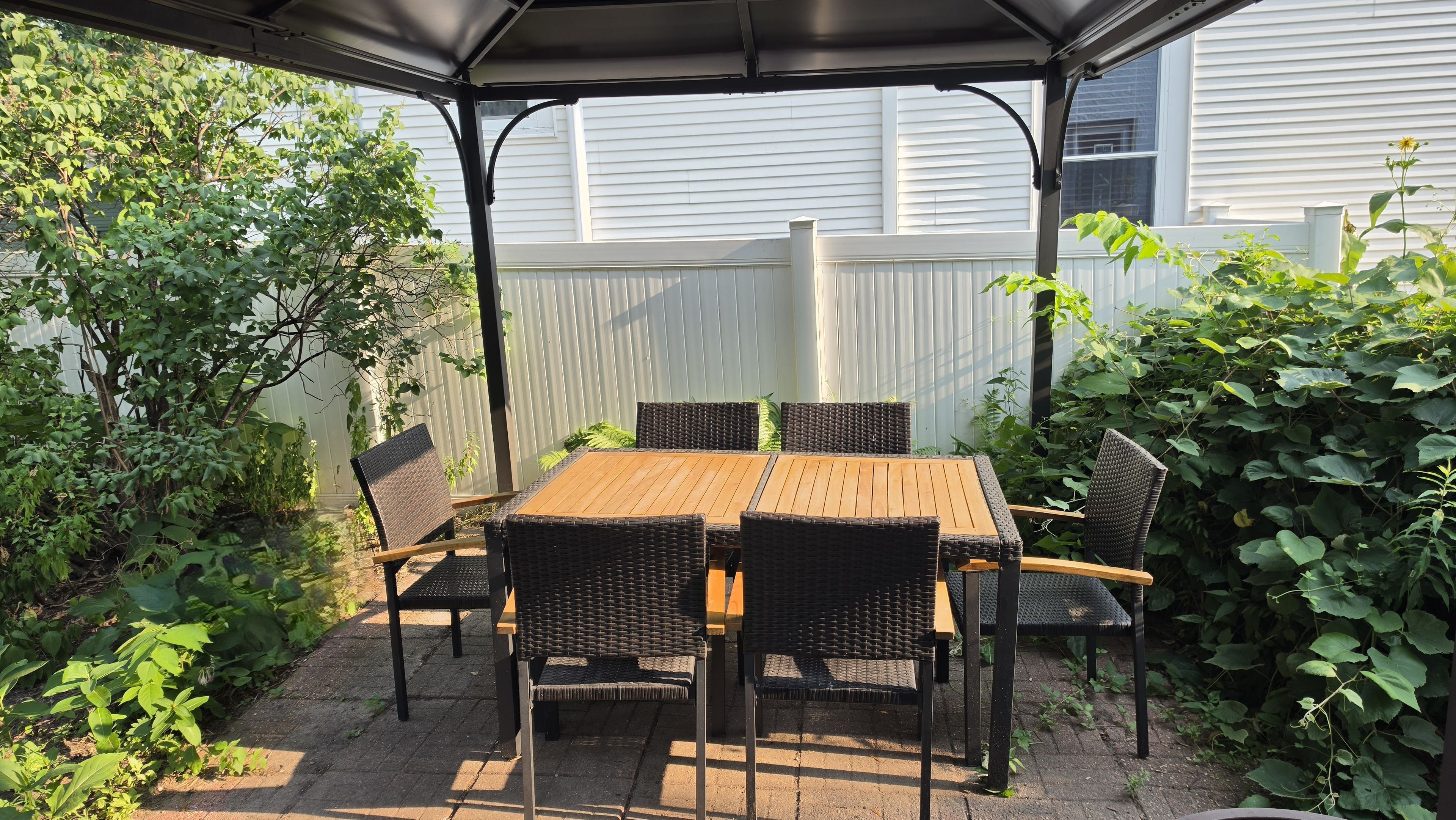 Un patio techado con una mesa de madera y sillas de mimbre, rodeado de follaje y una cerca blanca.
