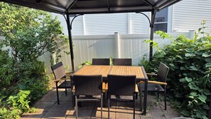 Une terrasse couverte avec une table en bois et des chaises en osier, entourée de verdure et d’une clôture blanche.