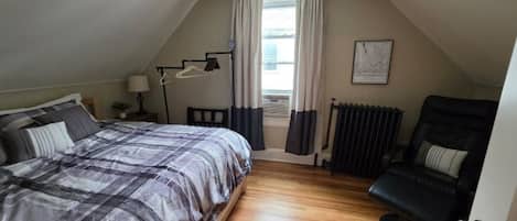 A bedroom with a bed, a chair, a radiator, and a window with curtains.