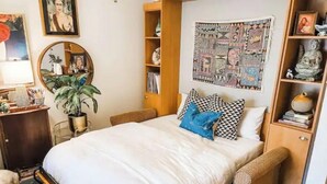 A bedroom with a bed, a bedside table, a chair, a shelf with decorative items, and a poster on the wall. - Cape Fear’s Original 1908 Cozy Historic Getaway (Wilmington)