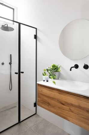 A modern bathroom with a glass shower enclosure, a wooden vanity, a round mirror, and a potted plant.