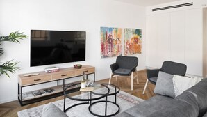 A modern living room with a flat-screen TV, a wooden console table, two armchairs, a round coffee table, and a grey sofa.