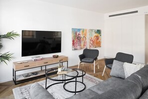A modern living room with a flat-screen TV, a wooden console table, two armchairs, a round coffee table, and a grey sofa.