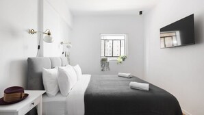 A modern bedroom with a bed, bedside table, lamp, window, and a flat-screen TV mounted on the wall.