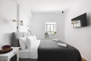 A modern bedroom with a bed, bedside table, lamp, window, and a flat-screen TV mounted on the wall.