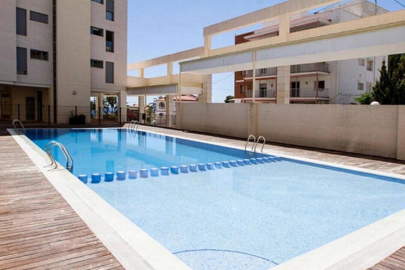 En rektangulär swimmingpool med ett trädäck och metallräcken, omgiven av en låg mur och angränsande byggnader.