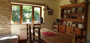 A dining area with a wooden table and chairs, a window with a view of greenery, a radiator, and a shelf with decorative plates and kitchenware. - Cottage on Lough Ree / River Shannon in the heart of Ireland (Newtowncashel)