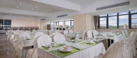 A large dining hall with tables set for service, a high ceiling, and large windows.