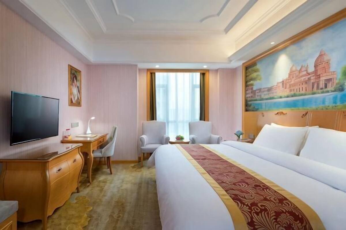 A hotel room with a bed, a desk, a chair, a television, and a painting on the wall.