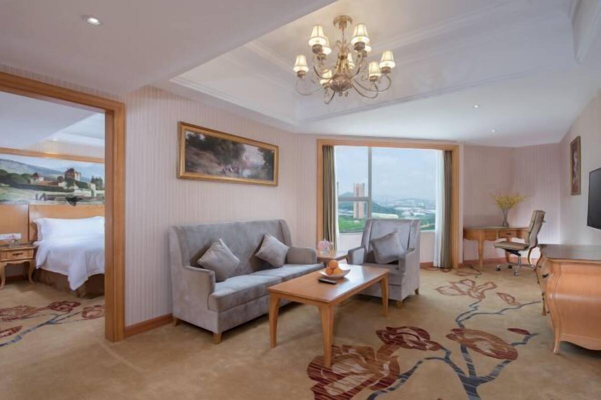A hotel room with a bed, a grey sofa, a wooden coffee table, a desk with a chair, a TV, and a large window with a view of a cityscape.