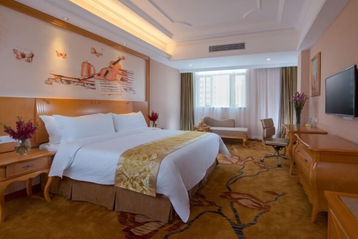 Une chambre d’hôtel avec un grand lit, un bureau, une chaise, une télévision et une fenêtre avec des rideaux.