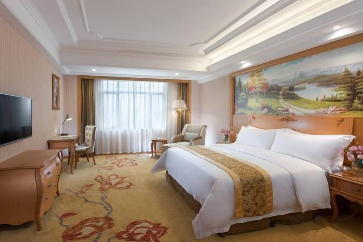A hotel room with a large bed, a desk, a chair, a television, and a painting on the wall.